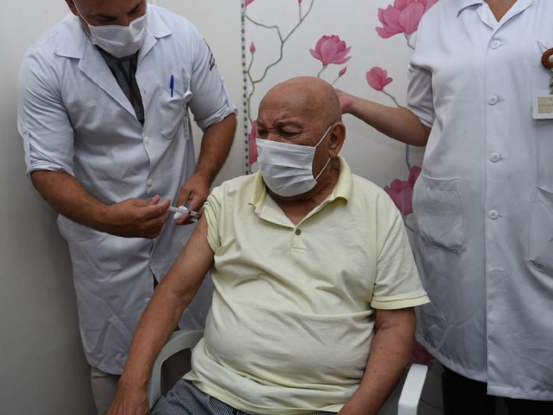 Um homem idoso recebe a vacina contra a covid no braço direito. Ele usa uma camiseta amarela e está de máscara. Do lado direito da foto está o enfermeiro que aplica a vacina e do lado direito uma enfermeira auxiliando. No fundo há um painel com flores rosa